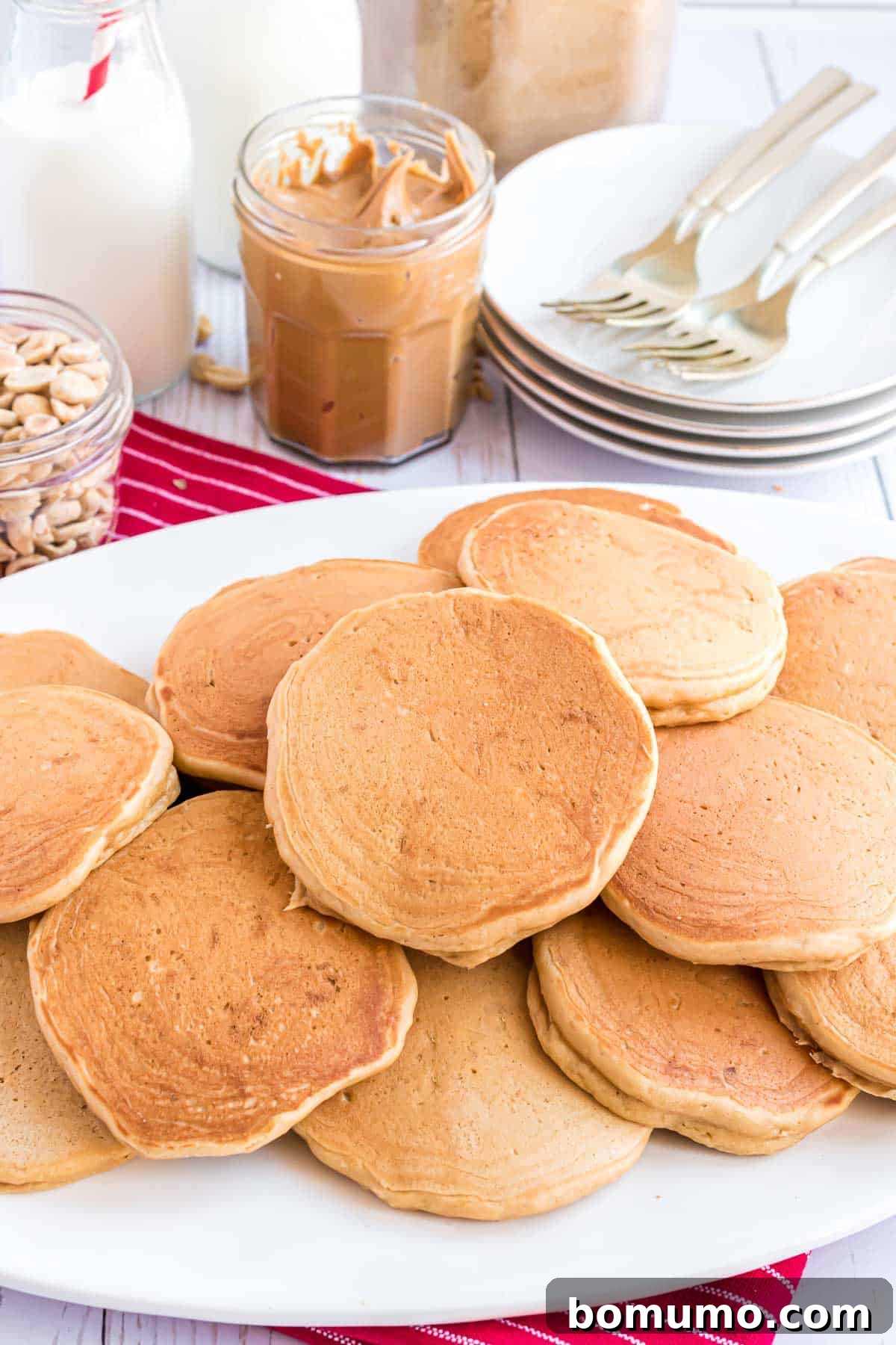 A bountiful pile of golden-brown pancakes rests invitingly on a pristine white plate, promising a delightful breakfast. In the background, elegant jars of creamy peanut butter and crunchy peanuts, alongside a neat stack of plates complete with forks, create a harmonious scene of culinary abundance.