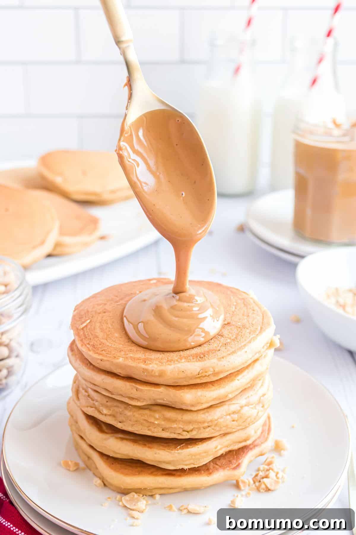 A luxurious spoon gracefully drizzles warm, molten peanut butter over a towering stack of perfectly cooked pancakes, artfully arranged on a pristine plate. In the softly blurred background, additional plates laden with more pancakes, a refreshing glass of milk, and charming jars of spreads complete this inviting breakfast tableau.