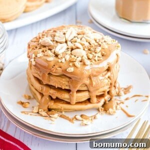 A glorious stack of fluffy peanut butter pancakes, generously topped with melted peanut butter and adorned with crunchy chopped peanuts, presented on a pristine white plate, ready to be enjoyed.