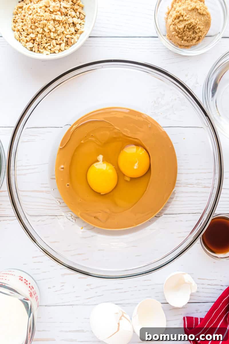 A gleaming glass bowl holds creamy peanut butter and two vibrant egg yolks, poised for whisking. Around it, smaller bowls contain meticulously chopped nuts, golden-brown sugar, fresh cream, and fragrant vanilla extract, all artfully arranged on a pristine white surface. Nearby, delicate eggshells and a charming red-striped cloth add a touch of rustic elegance to the scene.