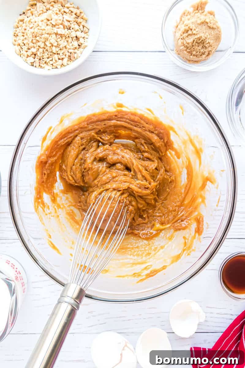 A pristine glass bowl showcases a creamy peanut butter and egg mixture, expertly blended with a whisk. Surrounding this central focus are artfully arranged small bowls containing finely chopped nuts, rich brown sugar, a precise measuring cup, delicate eggshells, and fragrant vanilla, all resting on a clean white surface, highlighting the meticulous preparation of ingredients.