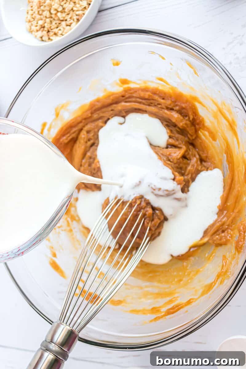 A skilled hand deftly whisks fresh milk into a luscious peanut butter mixture, gracefully swirling within a transparent glass bowl. In the softly blurred background, a small bowl of precisely chopped nuts awaits its turn, hinting at the delightful textures to come.