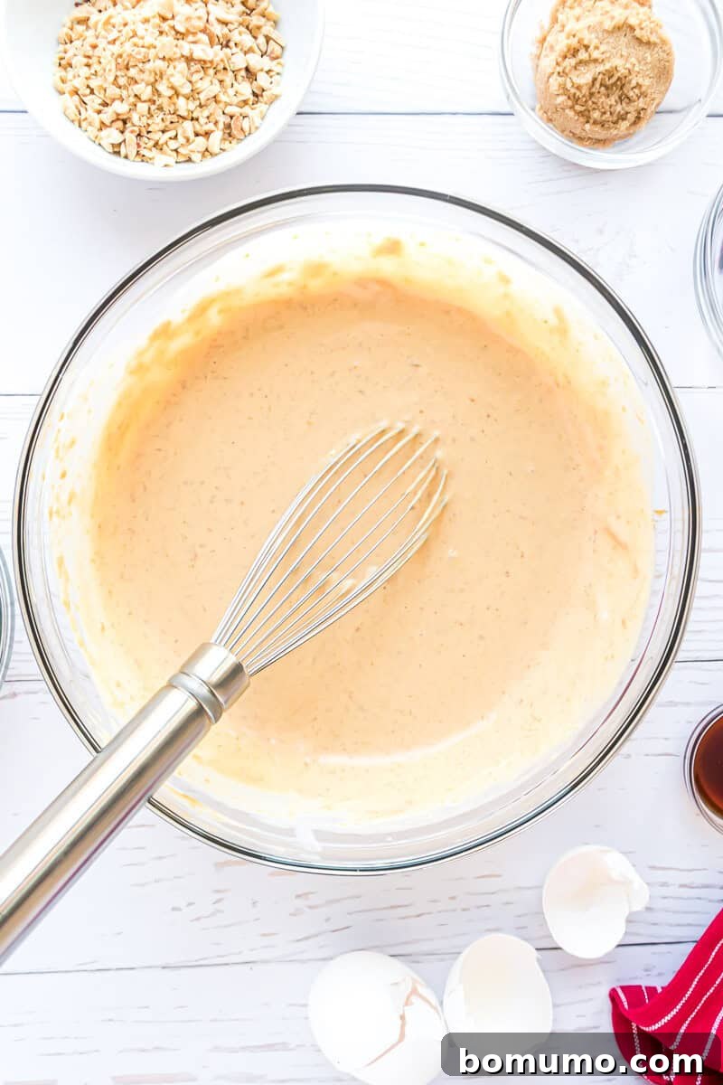 A vibrant mixing bowl contains a smooth, golden batter and a whisk, surrounded by an array of baking essentials: finely chopped nuts, rich brown sugar, a delicate bottle of vanilla extract, and neatly cracked eggshells, all elegantly arranged on a pristine white surface, ready for the next step in the baking process.