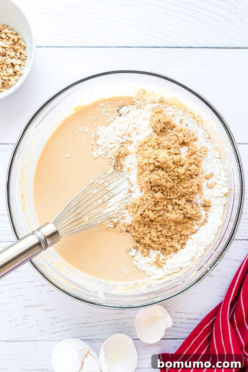 A pristine mixing bowl cradles a luscious batter, adorned with a sprinkle of flour and rich brown sugar, accompanied by a whisk ready for action. Nearby, delicate eggshells and a charming red-striped cloth rest gracefully on a clean white surface, capturing a moment of culinary creation.