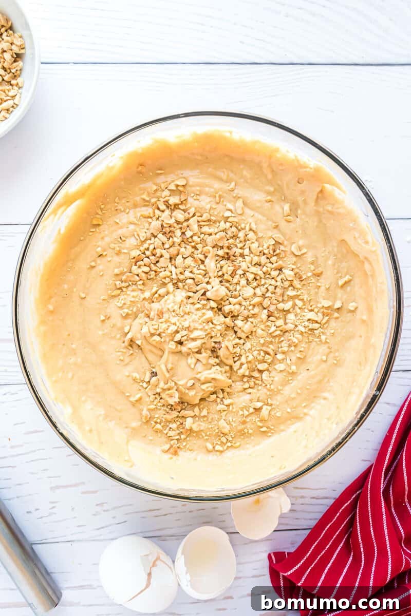 A clear glass bowl overflows with a rich, golden cake batter, generously topped with a sprinkle of crushed nuts, creating an inviting texture on a polished white wooden surface. Nearby, delicate cracked eggshells and a charming red-striped cloth add a touch of warmth and rustic appeal to the culinary setting.