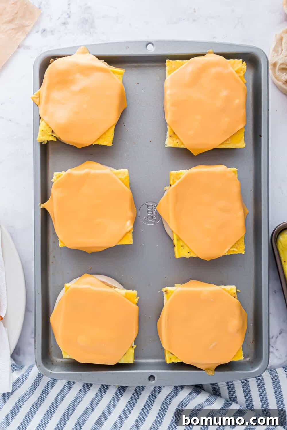 Breakfast Sandwich Bliss 11 Baking sheet with six pieces of english muffins, each topped with scrambled eggs and a slice of melted cheese.