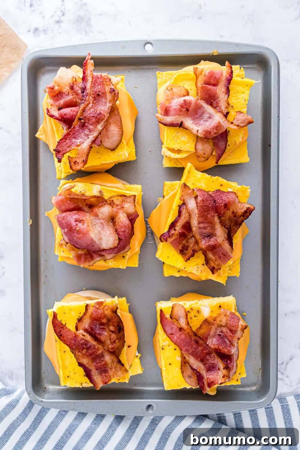 Breakfast Sandwich Bliss 12 A baking tray with six breakfast sandwiches, each with scrambled eggs, melted cheese, and crispy bacon on a english muffin.