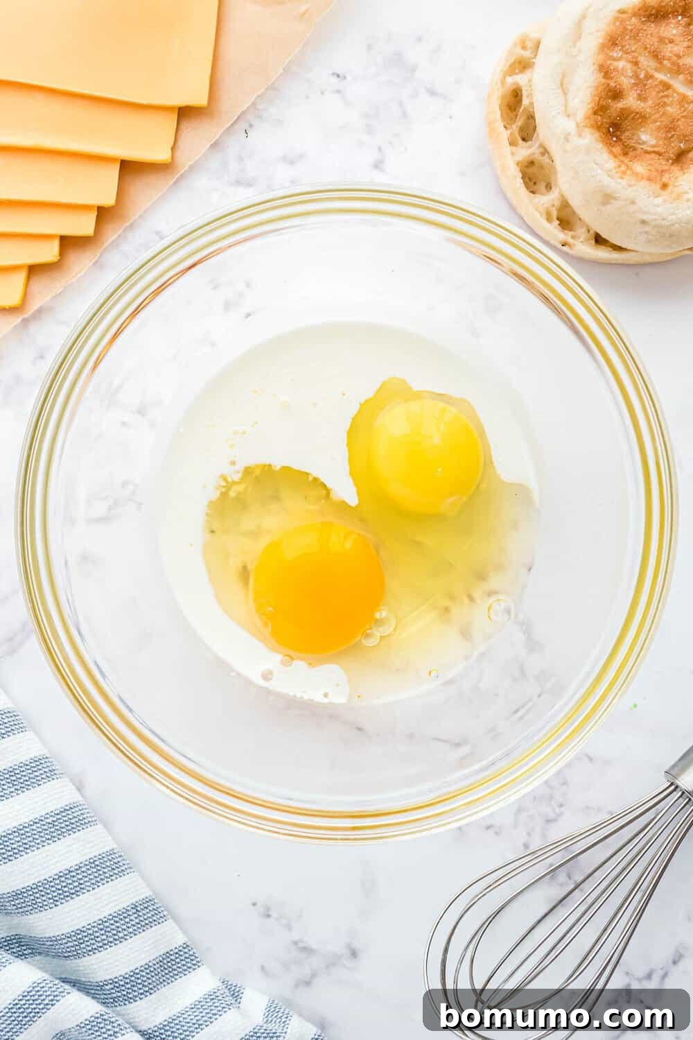 Breakfast Sandwich Bliss 14 Two cracked eggs in a glass bowl on a marble surface, with sliced cheese, an English muffin, a striped cloth, and a whisk nearby.
