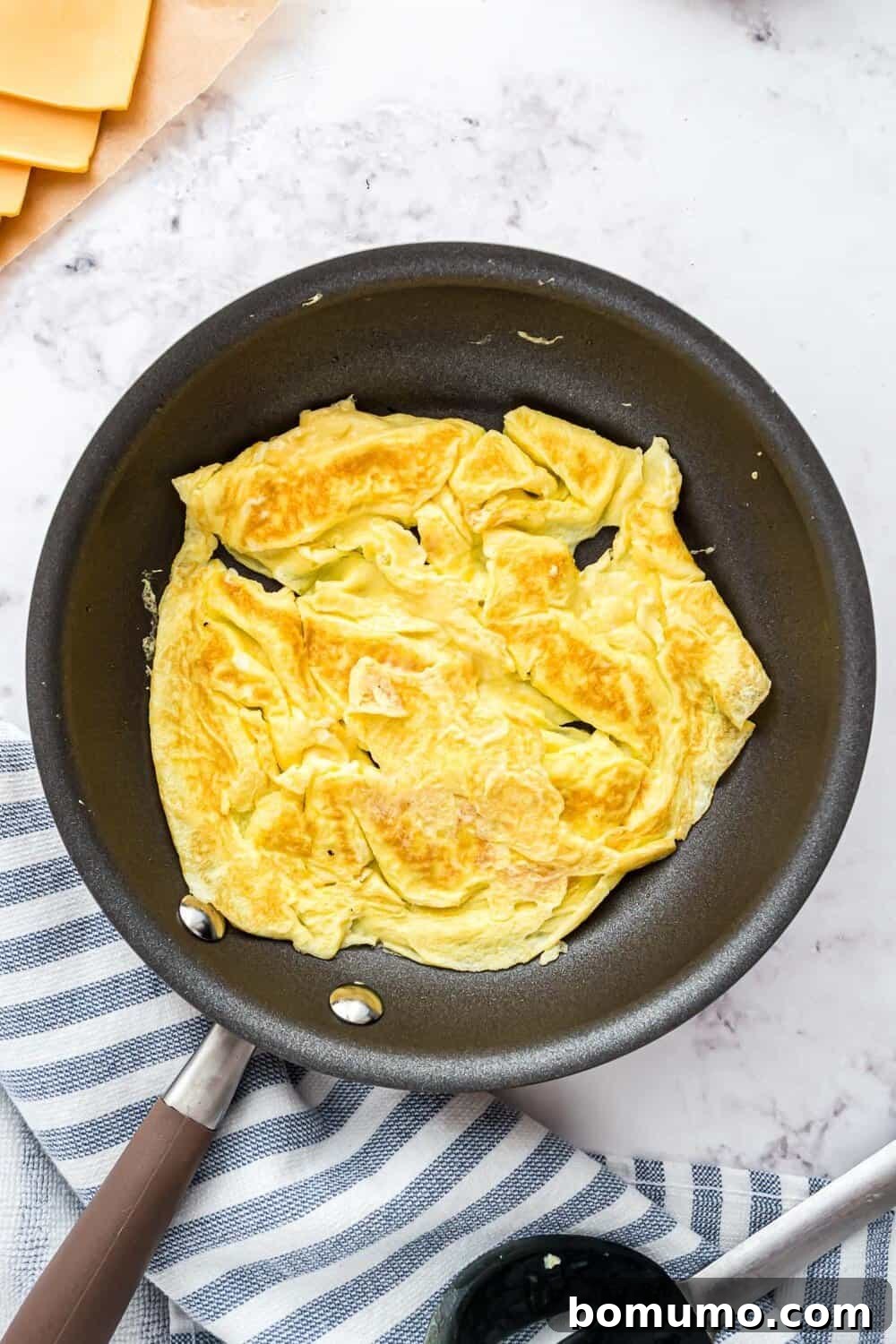 Breakfast Sandwich Bliss 15 Cooked eggs in a nonstick pan rests on a striped cloth on a marble countertop.