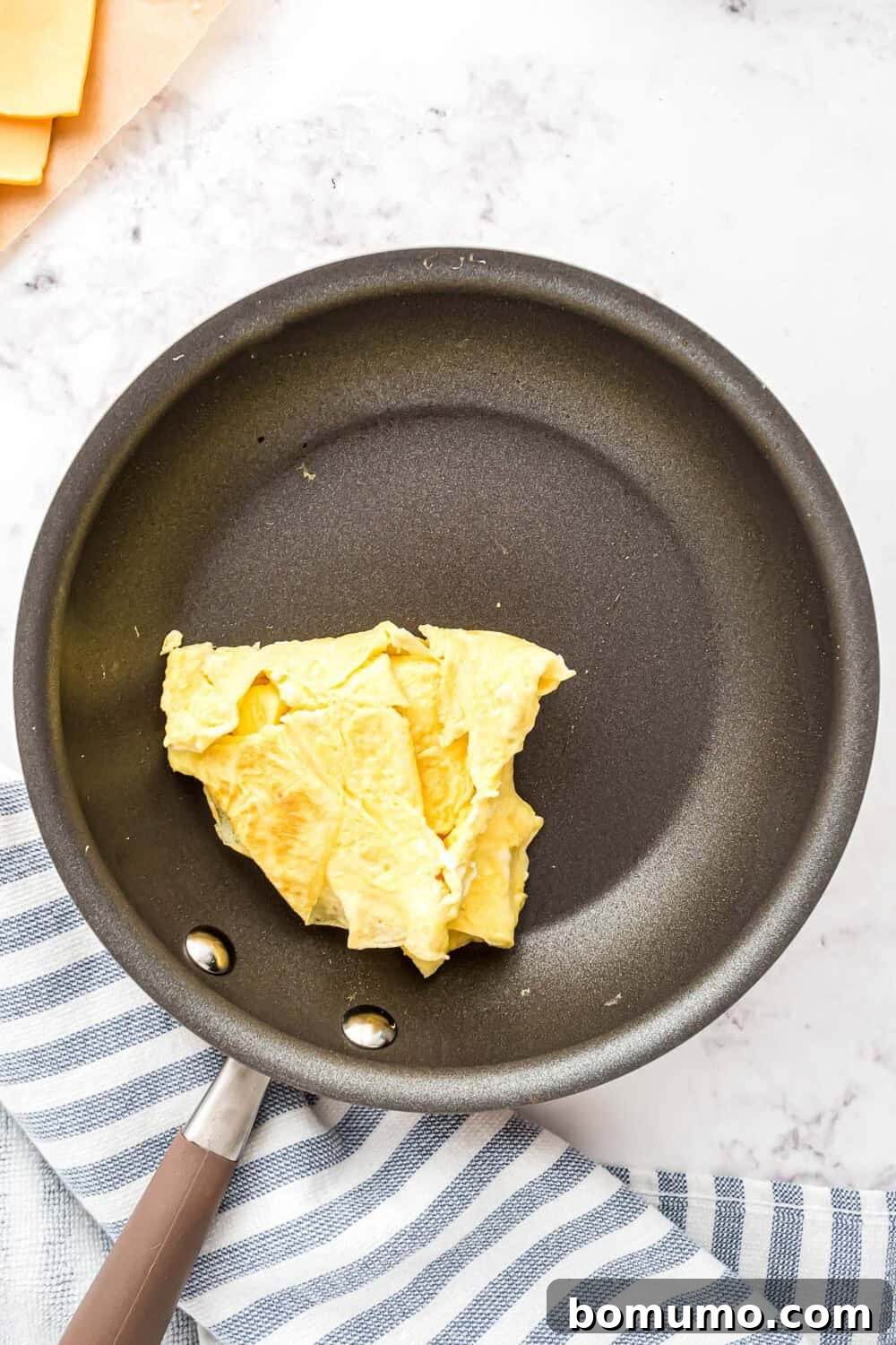 Breakfast Sandwich Bliss 17 A nonstick pan containing a folded scrambled egg, placed on a striped kitchen towel on a marble surface.