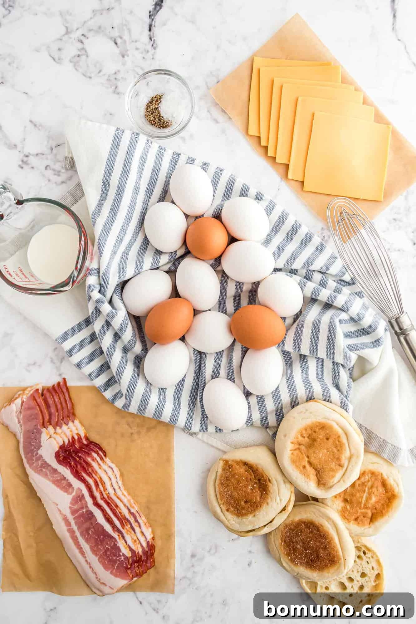 Breakfast Sandwich Bliss 4 Eggs on a striped cloth, bacon, cheese slices, English muffins, a whisk, a cup of milk, and a small bowl of salt and pepper on a marble surface.