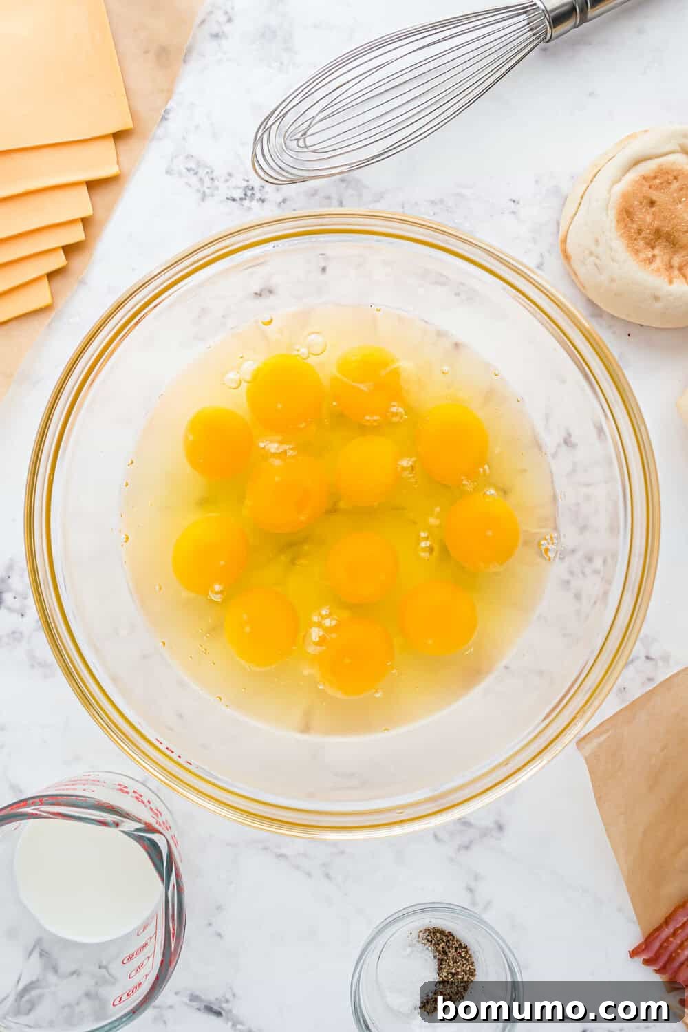 Breakfast Sandwich Bliss 5 A glass bowl with a dozen raw eggs on a marble countertop, surrounded by a whisk, cheese slices, a measuring cup with milk, salt, pepper, and an English muffin.