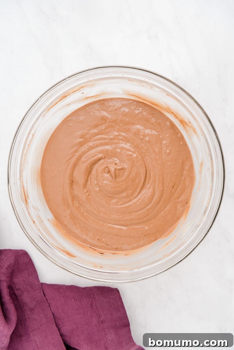 A large bowl of smooth, creamy chocolate pancake batter rests beside a vibrant purple cloth on a sleek marble surface, awaiting its delicious additions.
