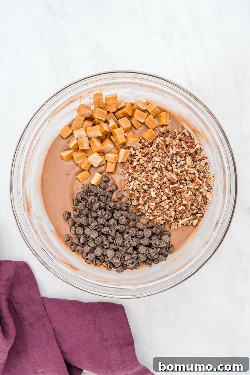 A mixing bowl filled with rich chocolate batter, generously topped with chocolate chips, cubed caramel pieces, and crunchy chopped pecans, sits enticingly on a marble surface with a purple cloth nearby.