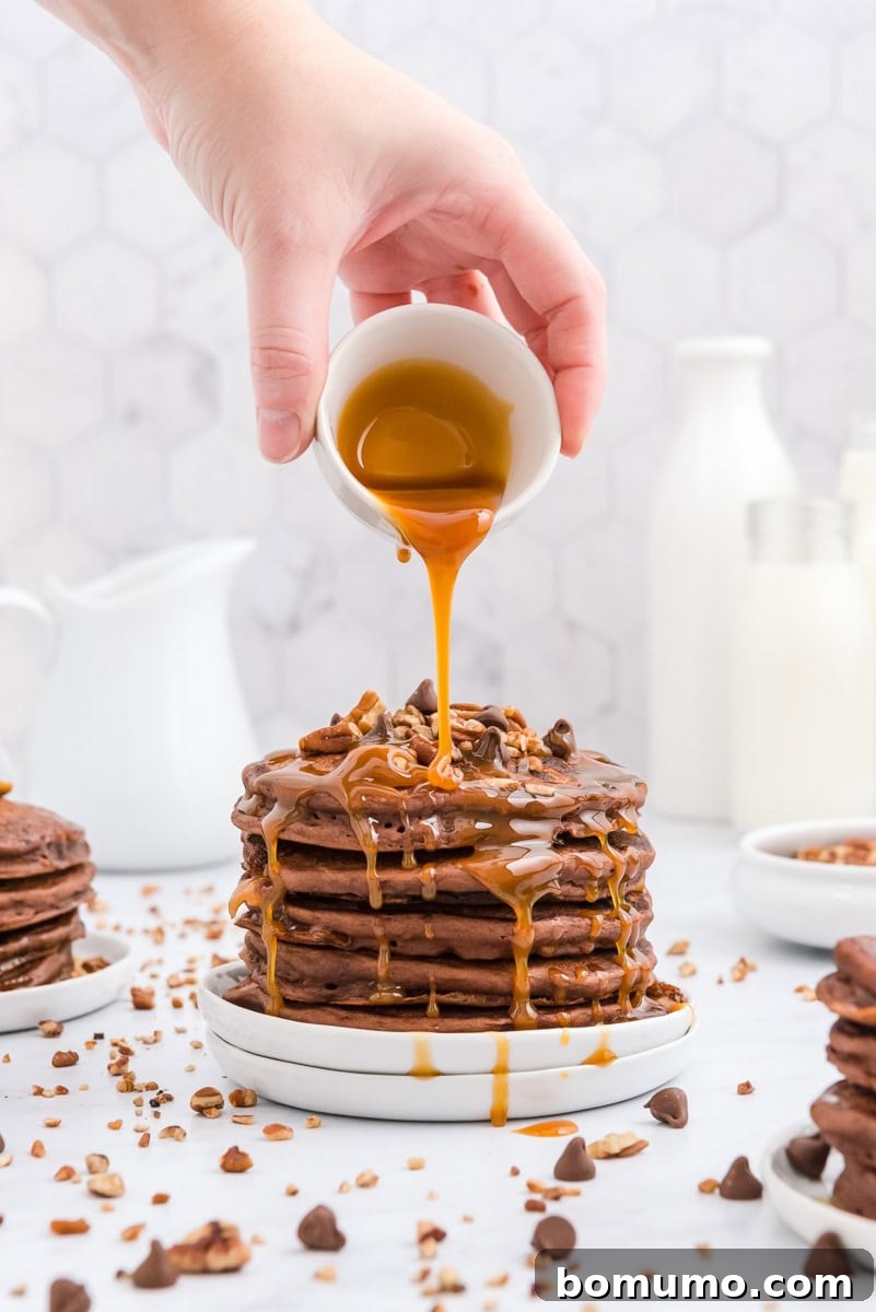 A hand gently pours rich syrup over a towering stack of delectable chocolate pancakes, generously adorned with crunchy pecan pieces and sweet chocolate chips, elegantly presented on a white plate.