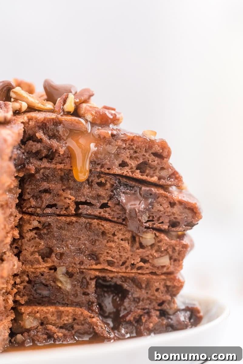 A tantalizing close-up of a stack of chocolate pancakes, revealing perfectly melted chocolate chips and visible pecan pieces, with luscious gooey caramel syrup cascading down the sides.