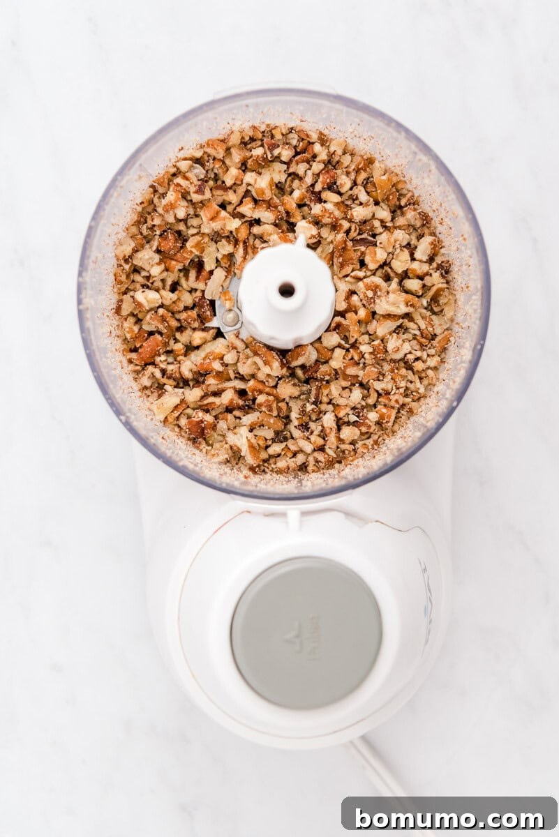 A clear glass bowl filled with roughly chopped pecans, ready for toasting, sits on a pristine white marble surface.