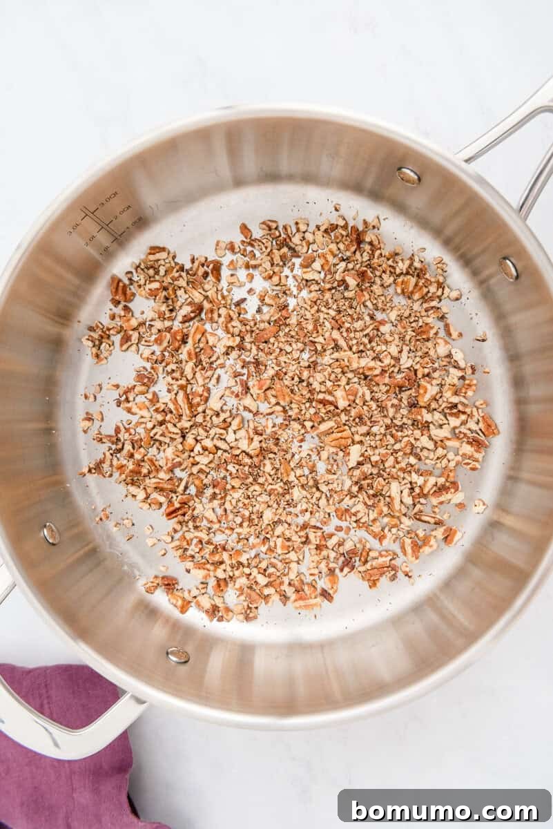 Chopped pecans are actively being toasted in a gleaming stainless steel pan, resting on a cool marble surface, emitting a rich, nutty aroma.