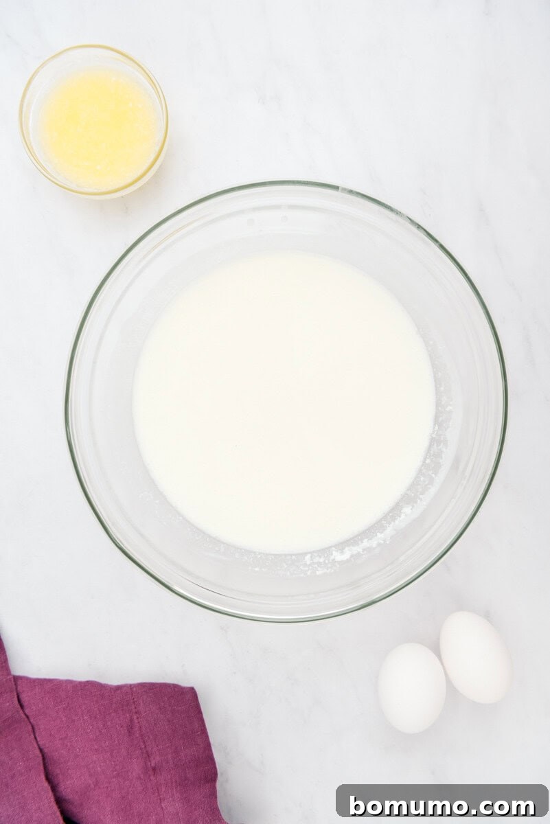 A glass bowl holds fresh milk, accompanied by a small dish of golden melted butter, two pristine eggs, and a soft purple cloth, all set on a clean white surface.