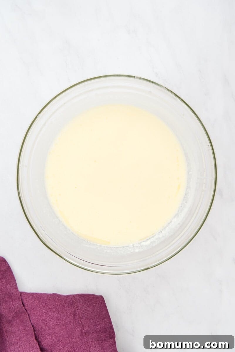A pristine glass bowl containing perfectly combined wet ingredients (milk, melted butter, and eggs) rests on a smooth marble surface, with a hint of purple cloth visible nearby.