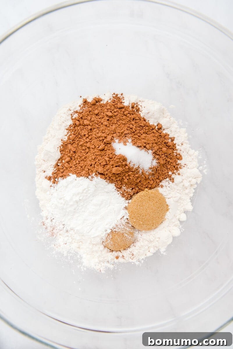 A gleaming glass bowl on a polished marble surface holds a carefully measured blend of dry pancake ingredients: flour, cocoa powder, baking powder, brown sugar, and salt.