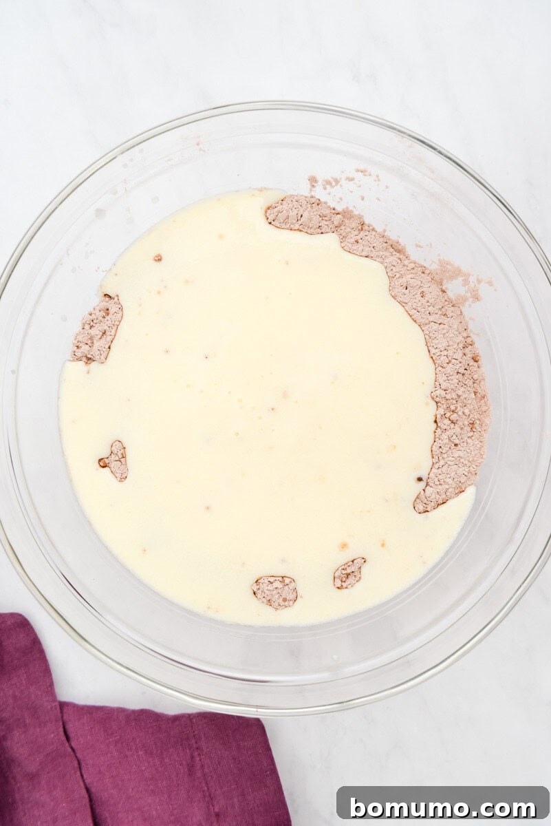A clear glass bowl contains a perfectly blended mixture of liquid and dry pancake ingredients, resulting in a smooth, thick chocolate batter. A soft purple cloth is subtly placed beneath the bowl.