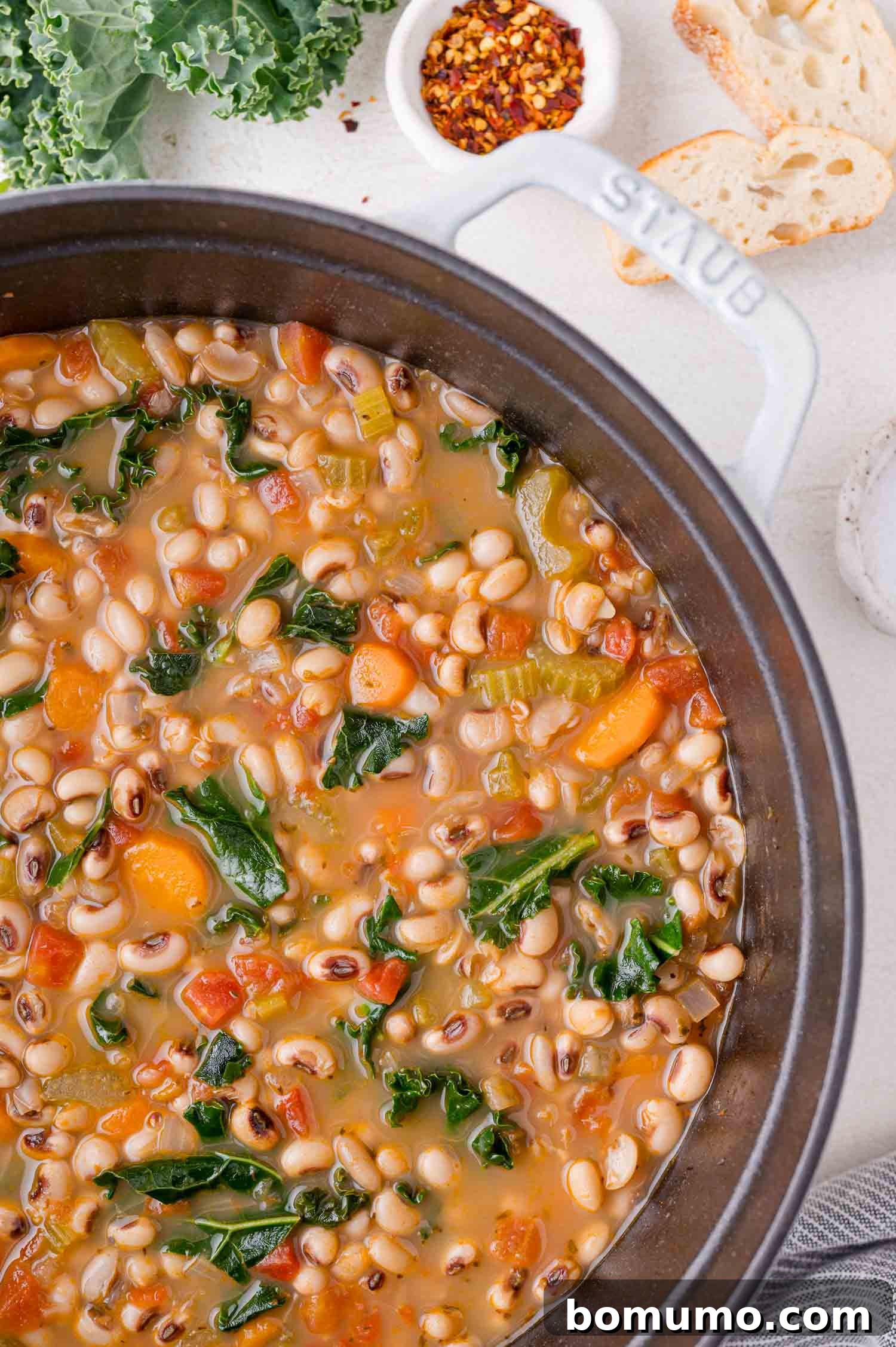 A pot of black-eyed pea soup with carrots, kale, and white beans, surrounded by slices of bread and a small bowl of red pepper flakes.