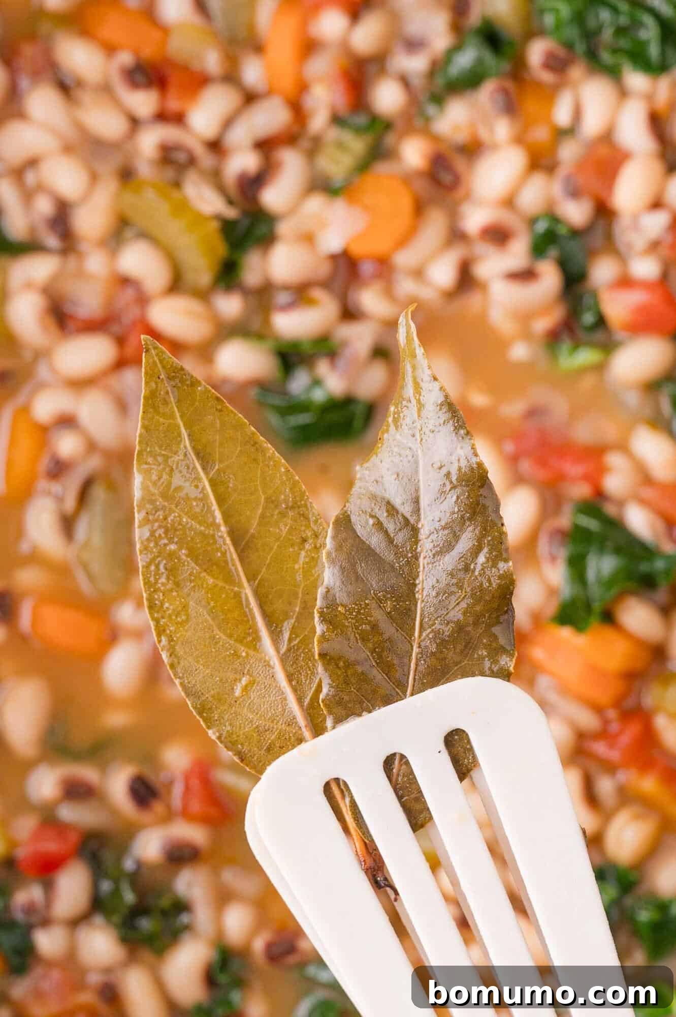 Two bay leaves held on a slotted spatula above a pot of vegetable and black-eyed pea soup.