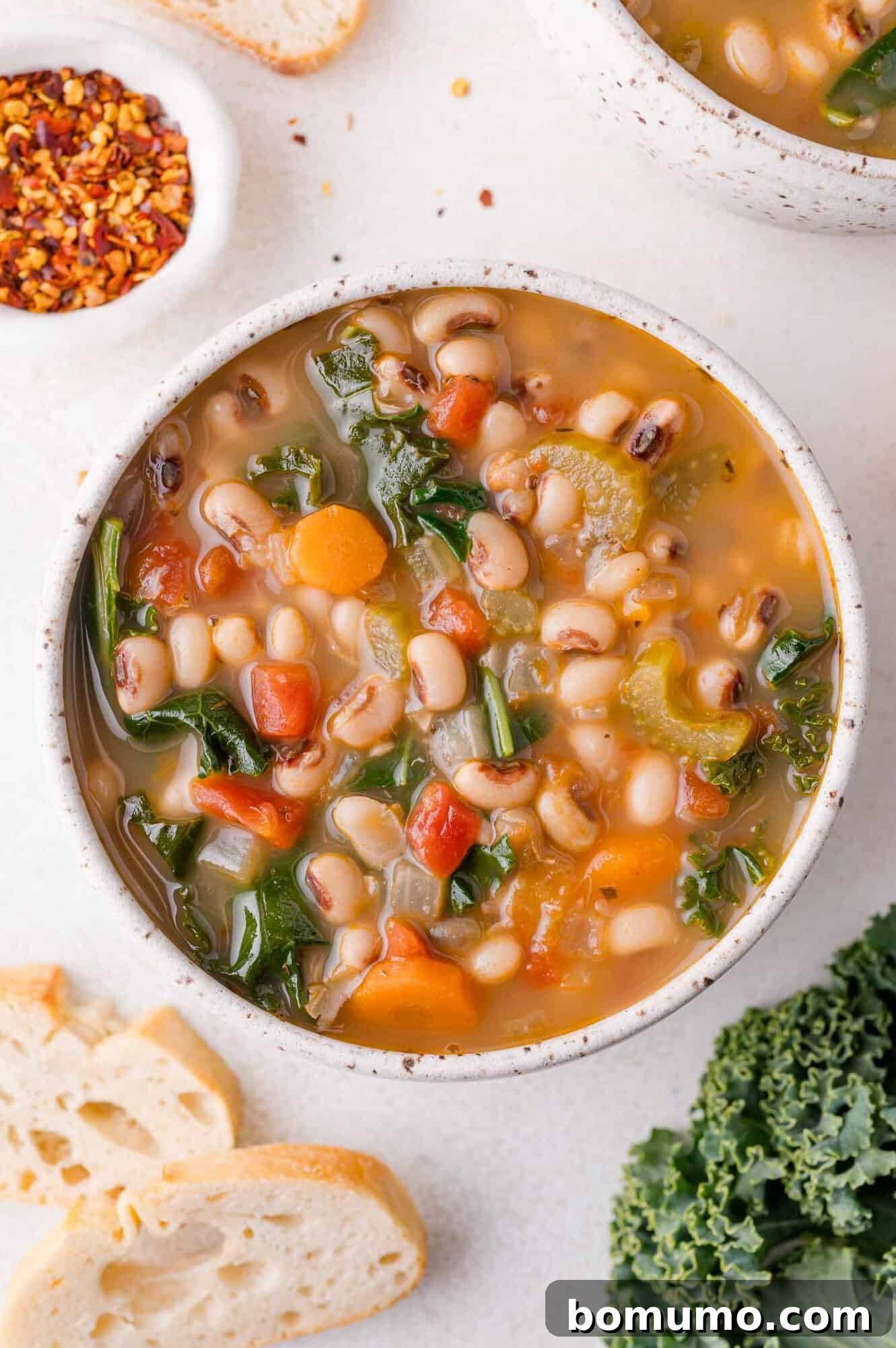 A bowl of black-eyed pea soup with tomatoes, greens, and carrots, accompanied by slices of bread and a small dish of red pepper flakes.