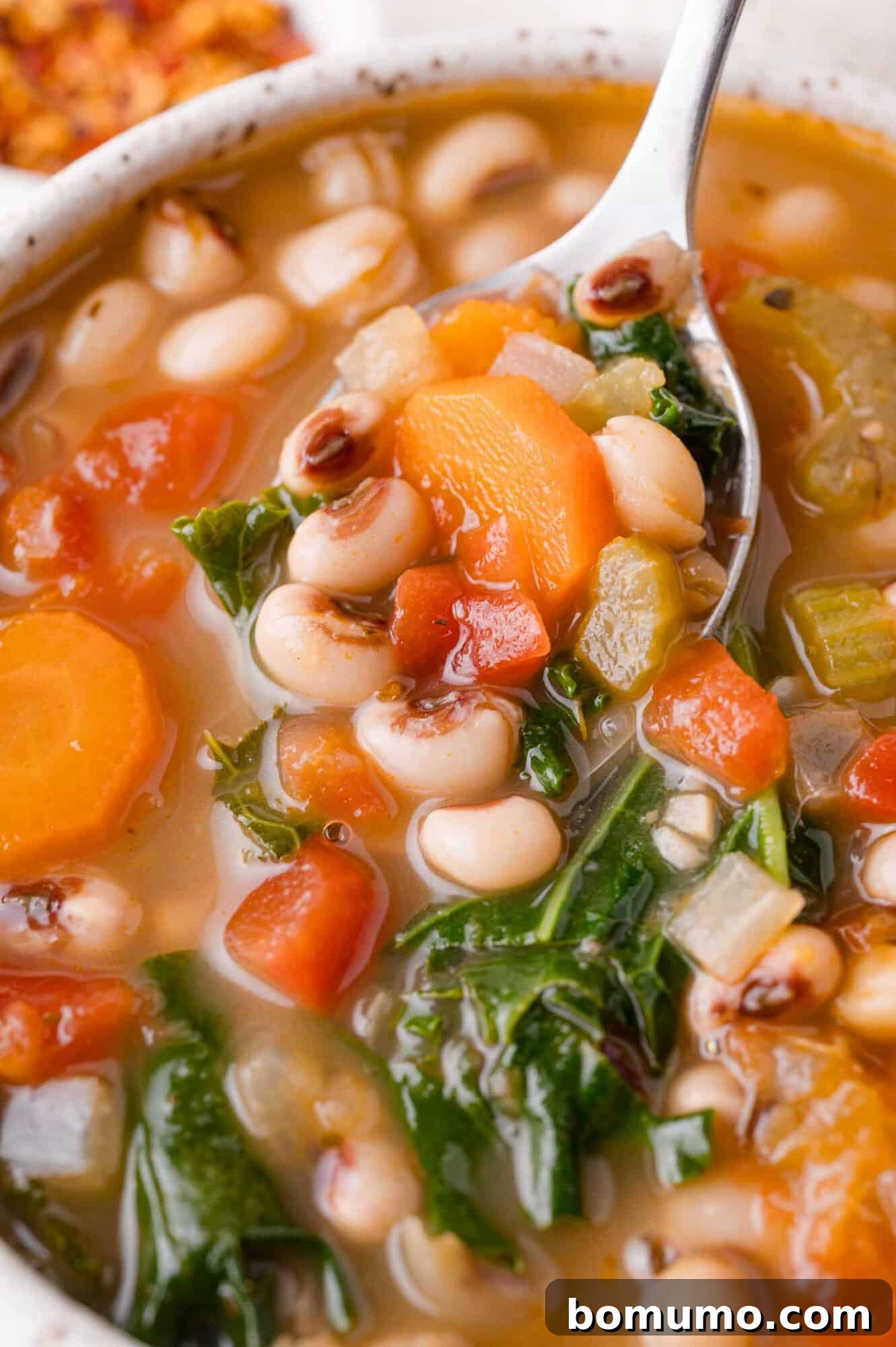 A close-up of a bowl of black-eyed pea soup with carrots, tomatoes, celery, and leafy greens, with a spoon lifting a portion.