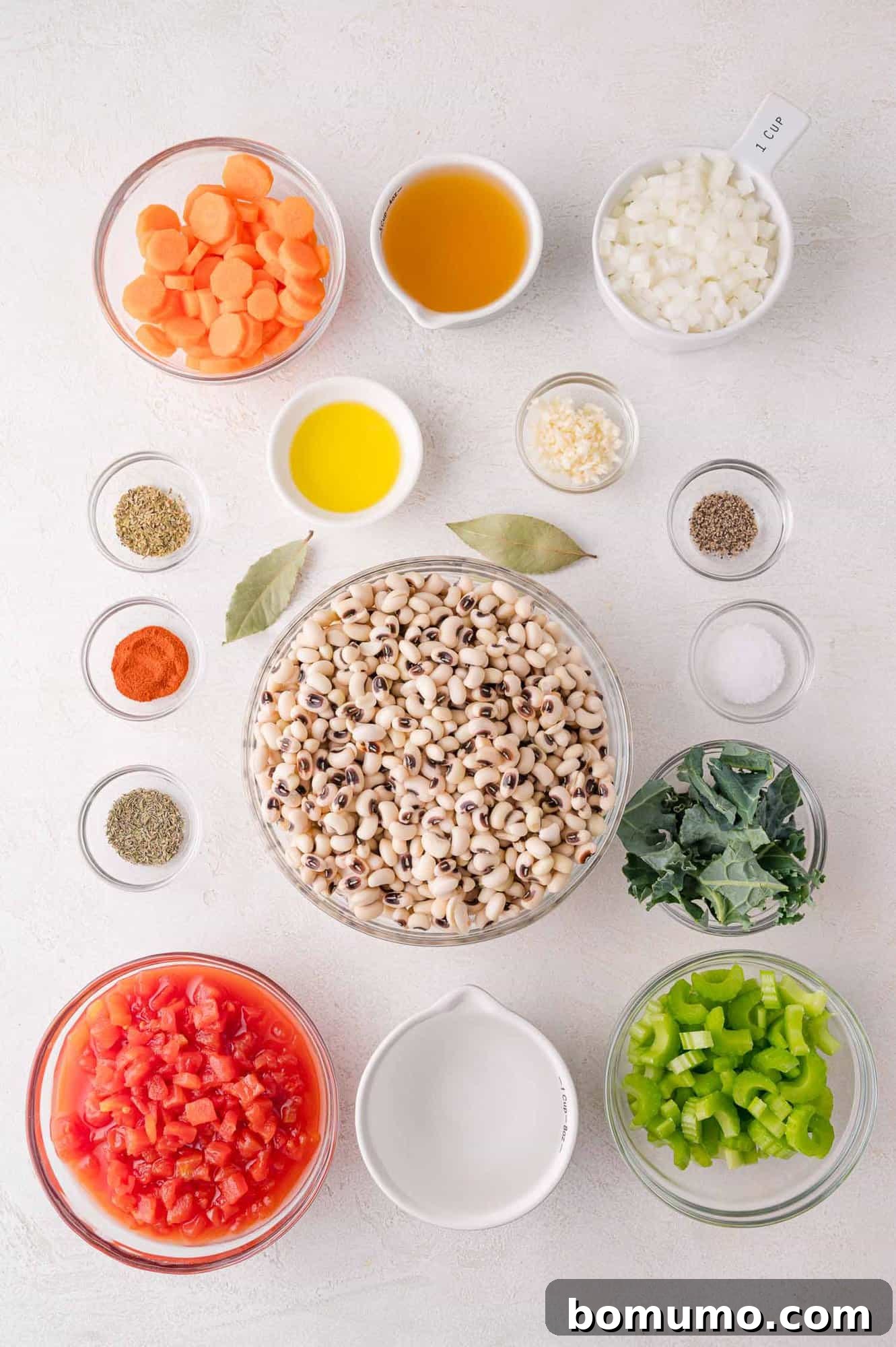 Overhead view of prepped ingredients in bowls, including black-eyed peas, diced tomatoes, carrots, celery, onion, garlic, kale, spices, bay leaves, broth, and oil on a white surface.