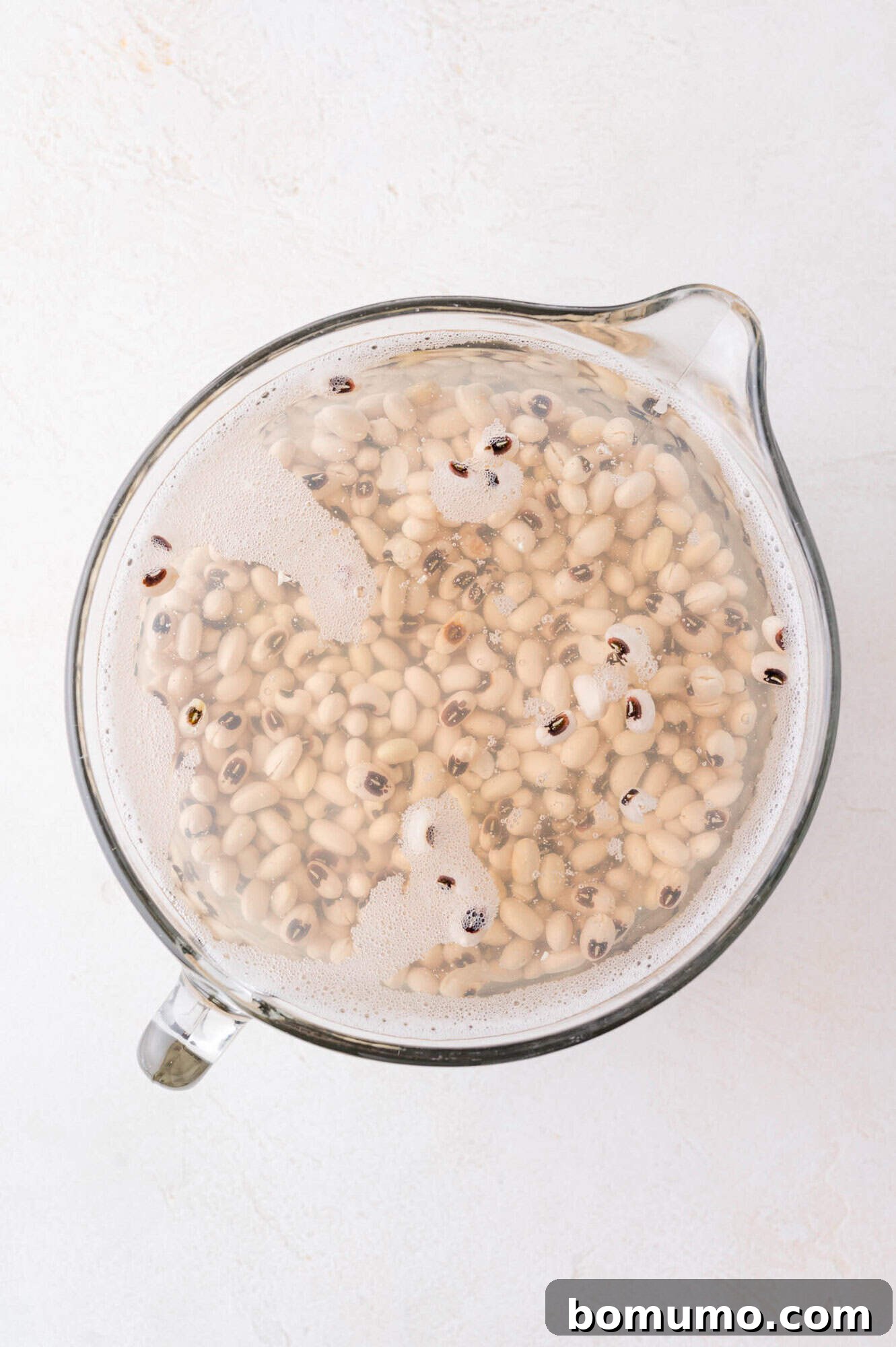 A glass measuring cup filled with water and soaking black-eyed peas, seen from above on a white surface.