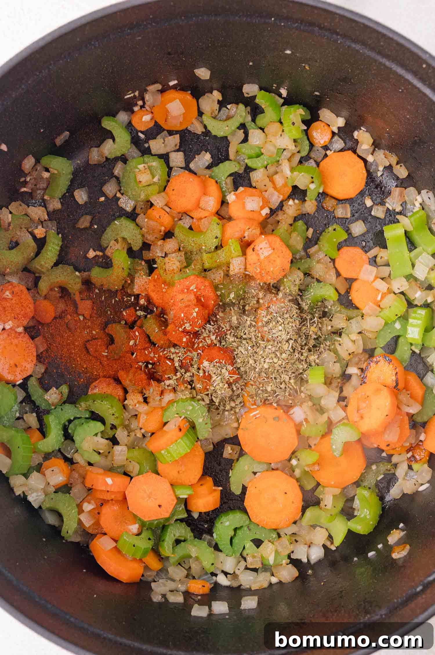 Chopped carrots, celery, and onions sautéing in a black pot with dried herbs and spices sprinkled on top.