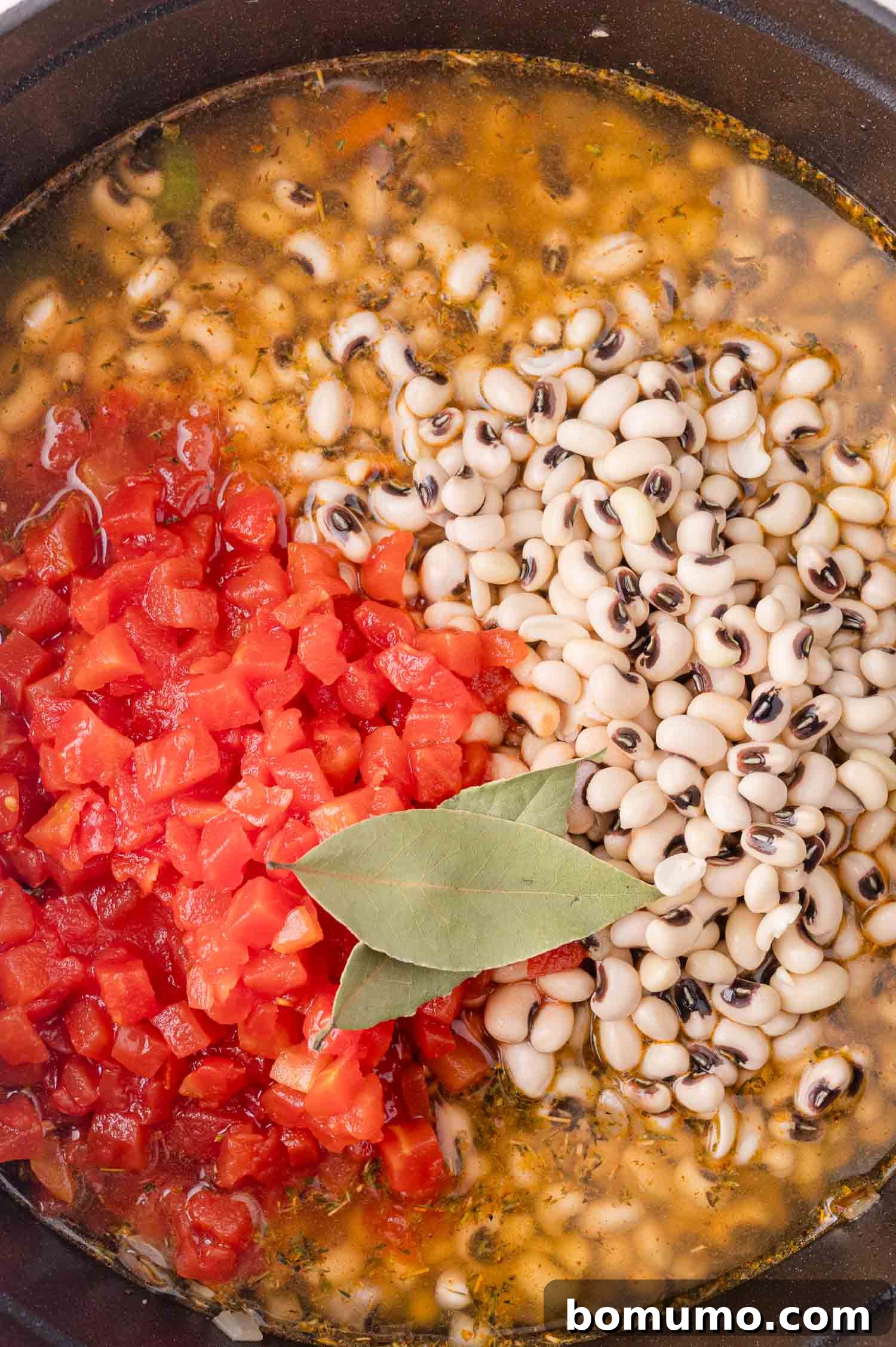A pot containing black-eyed peas, diced tomatoes, seasonings, and two bay leaves in a simmering broth.