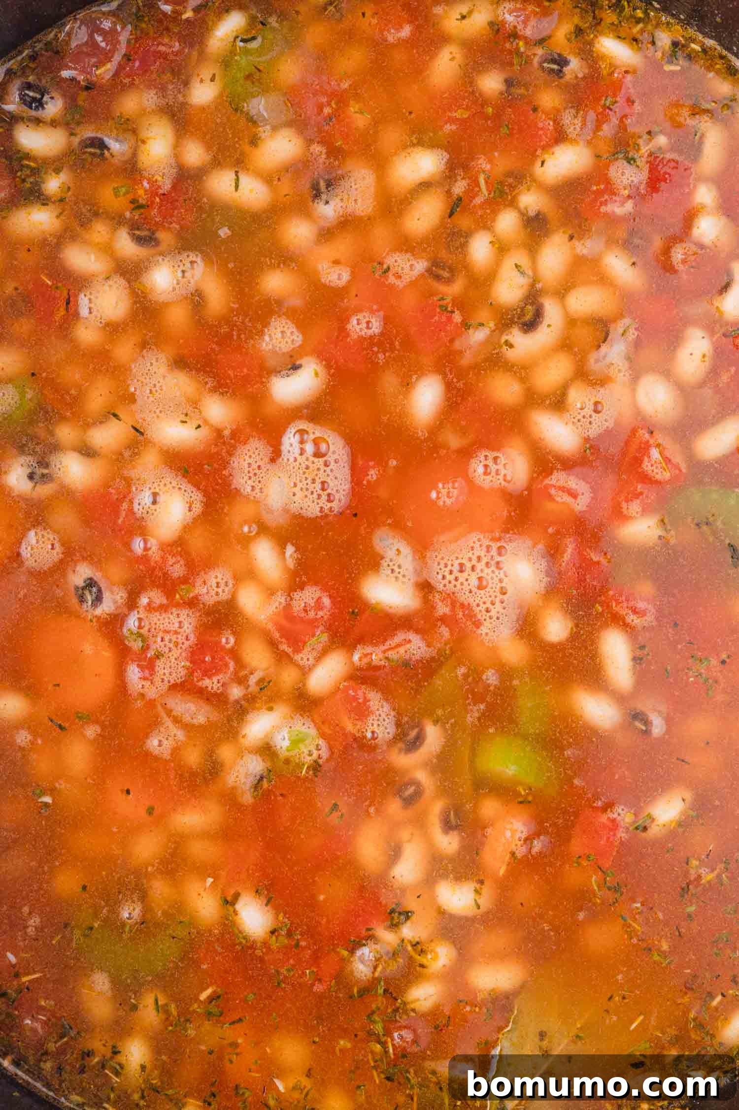A close-up of a pot of stew with black-eyed peas, diced tomatoes, celery, and herbs in a red broth.