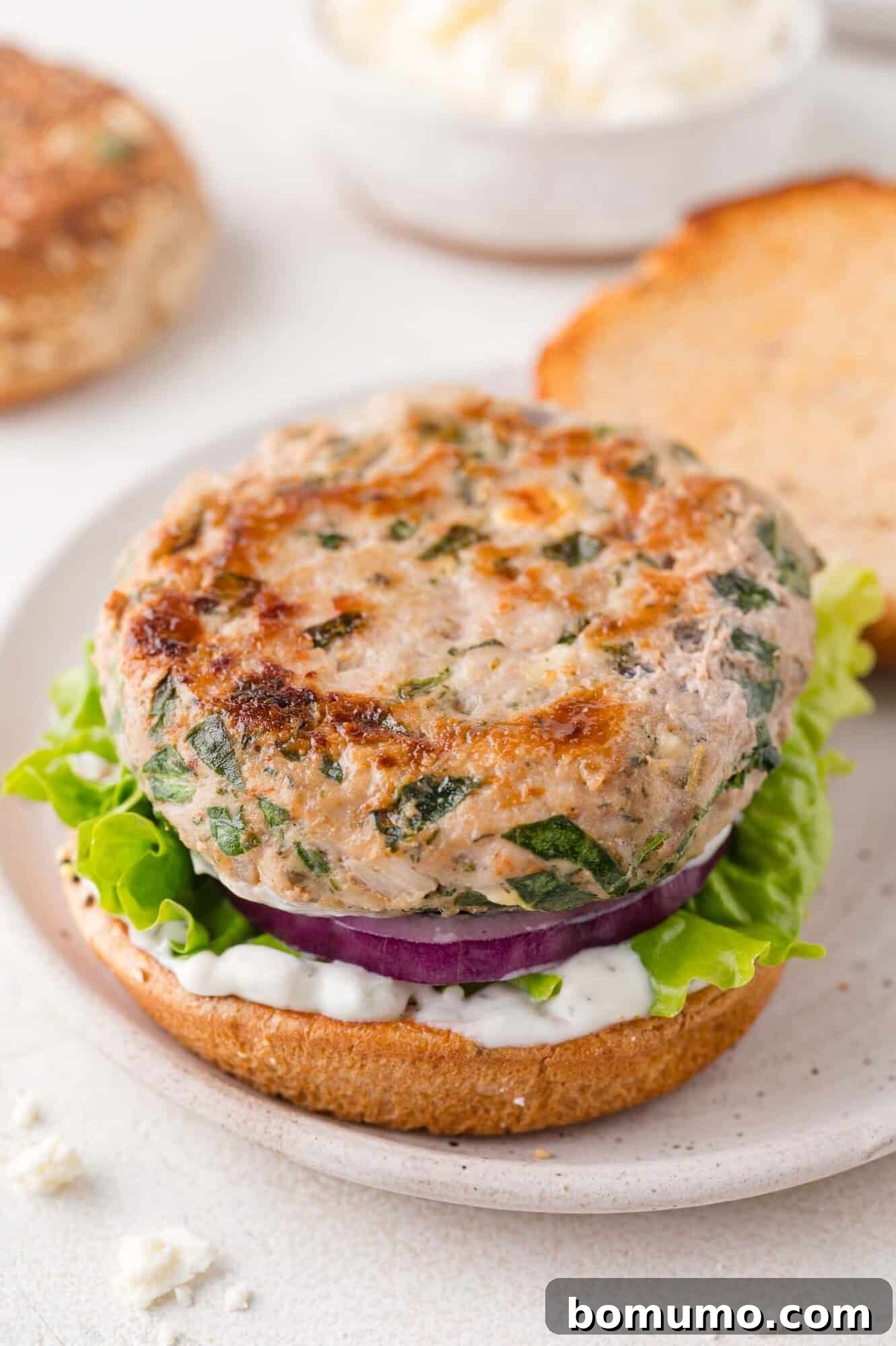 An open-faced Greek turkey burger with fresh spinach, crisp lettuce, thinly sliced red onion, and creamy tzatziki sauce on a toasted bun, presented on a pristine white plate.