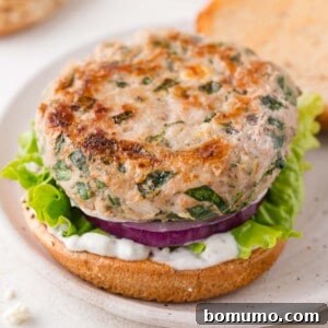 A turkey burger patty with herbs on a bun, layered with green leaf lettuce, sliced red onion, and white sauce, served open-faced on a plate.