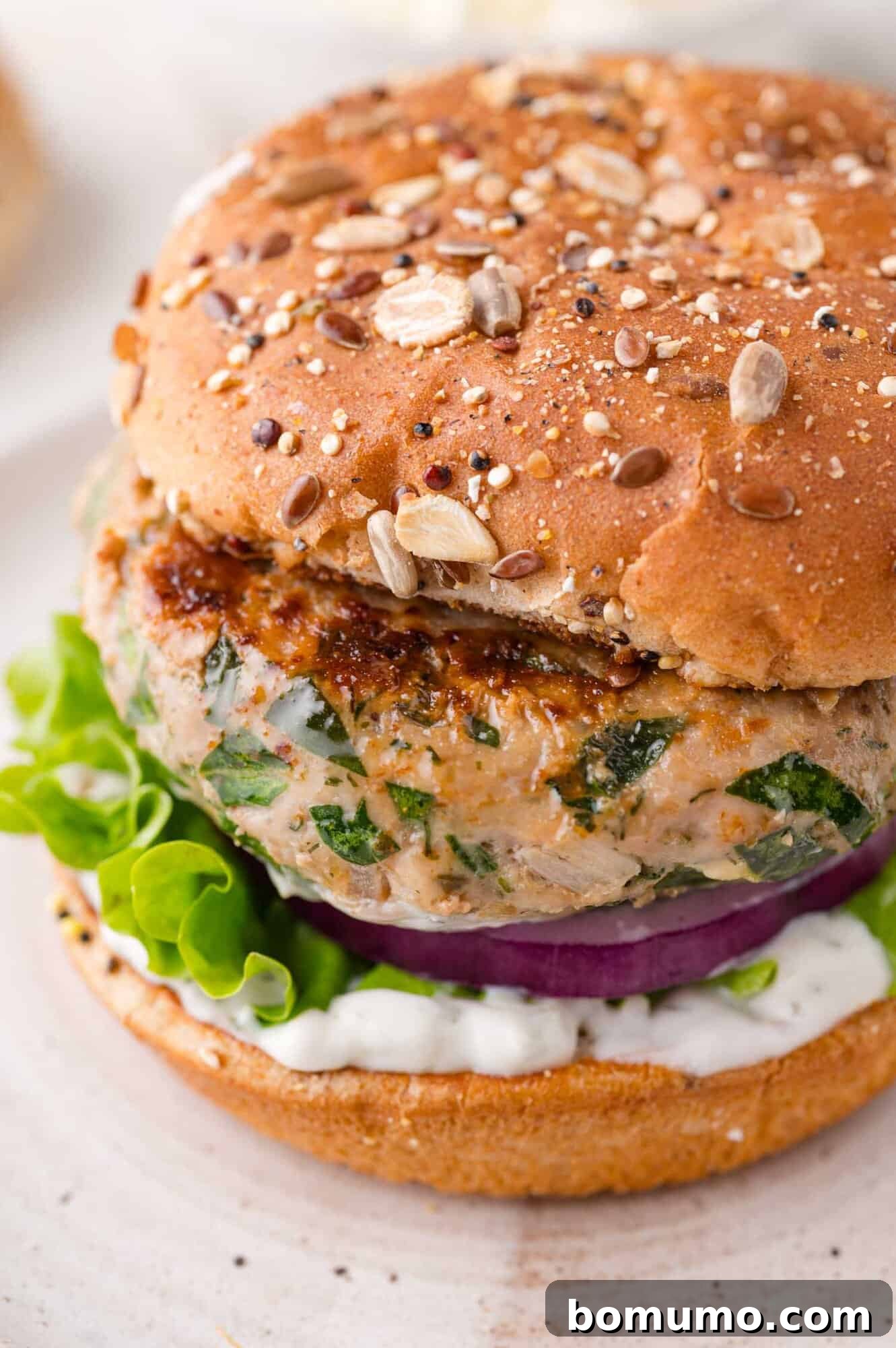 A close-up of a perfectly cooked Greek turkey burger resting on a whole-grain bun, beautifully garnished with crisp lettuce, thin slices of red onion, and a generous spread of creamy white tzatziki sauce.