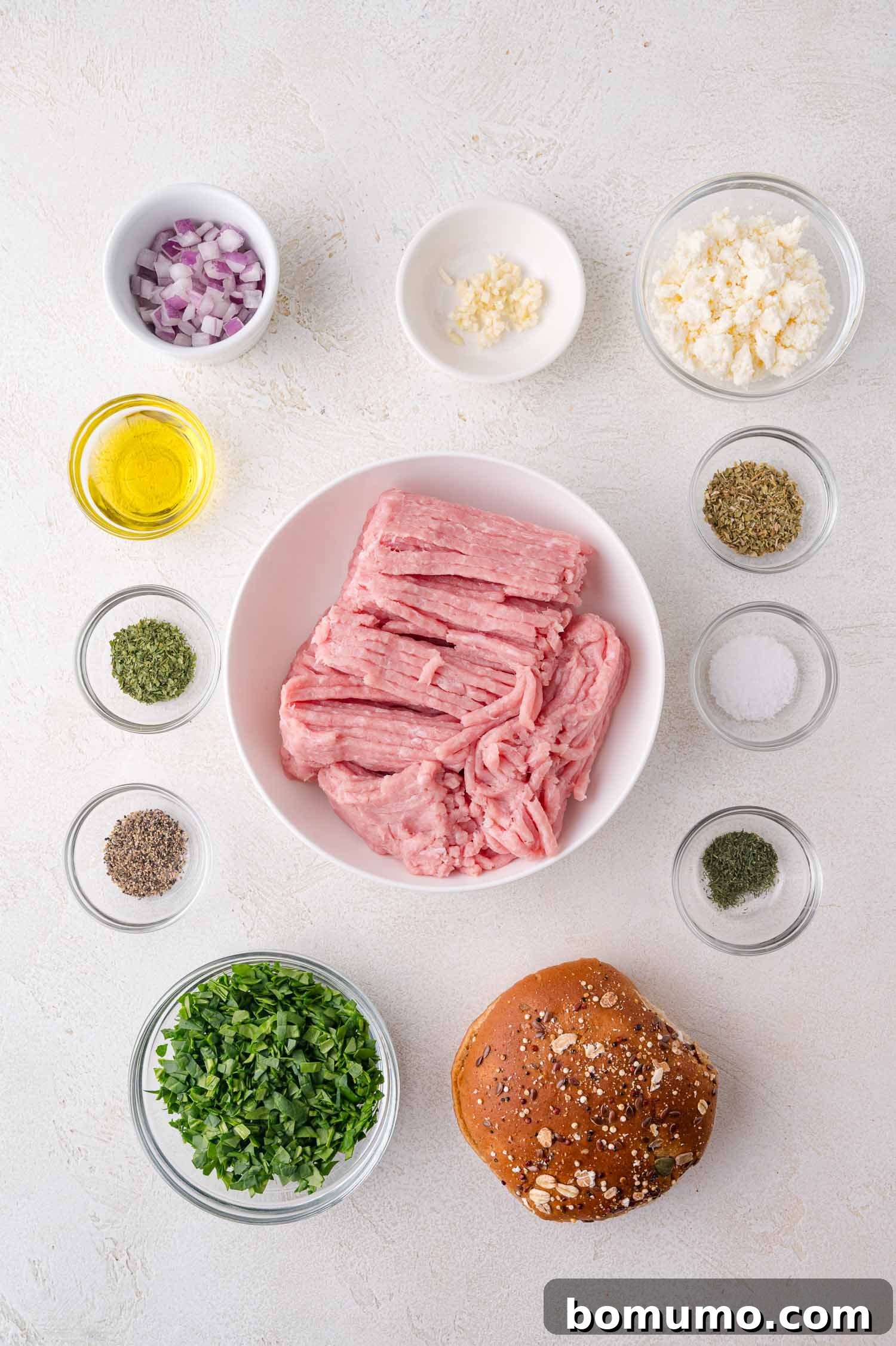 Ground turkey in a bowl surrounded by small bowls of chopped onion, garlic, parsley, feta, spices, olive oil, and an oat-topped burger bun on a white surface.