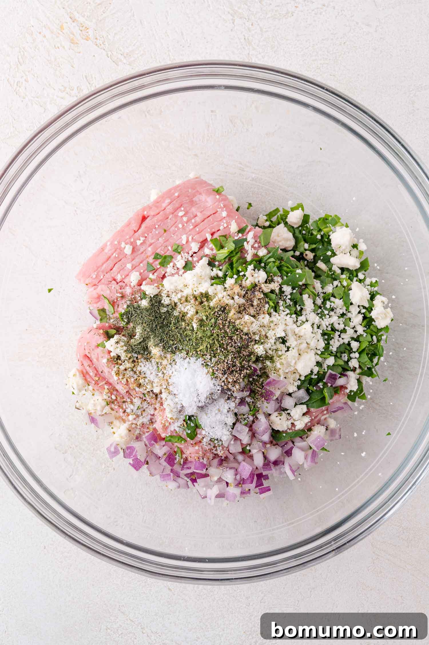 Ground meat, chopped red onions, herbs, spices, salt, and crumbled cheese are arranged in a clear glass mixing bowl on a light surface.