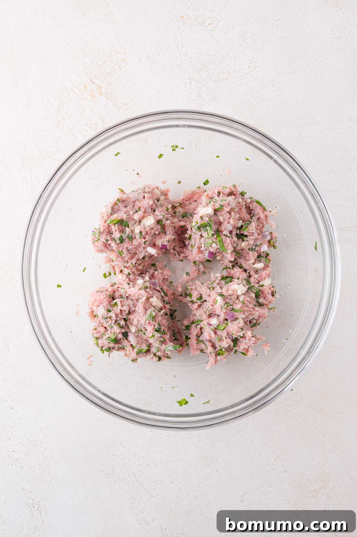 A glass bowl containing a mixture of ground meat, herbs, and chopped onions divided into four portions on a light surface.