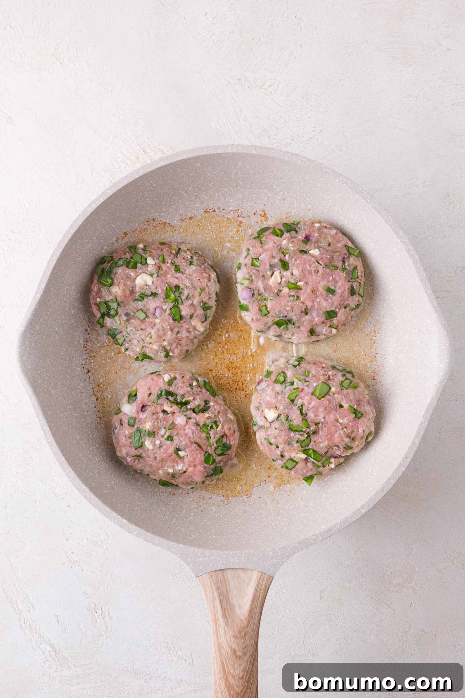 Four raw patties with herbs and diced vegetables are cooking in a light-colored skillet on a white surface.