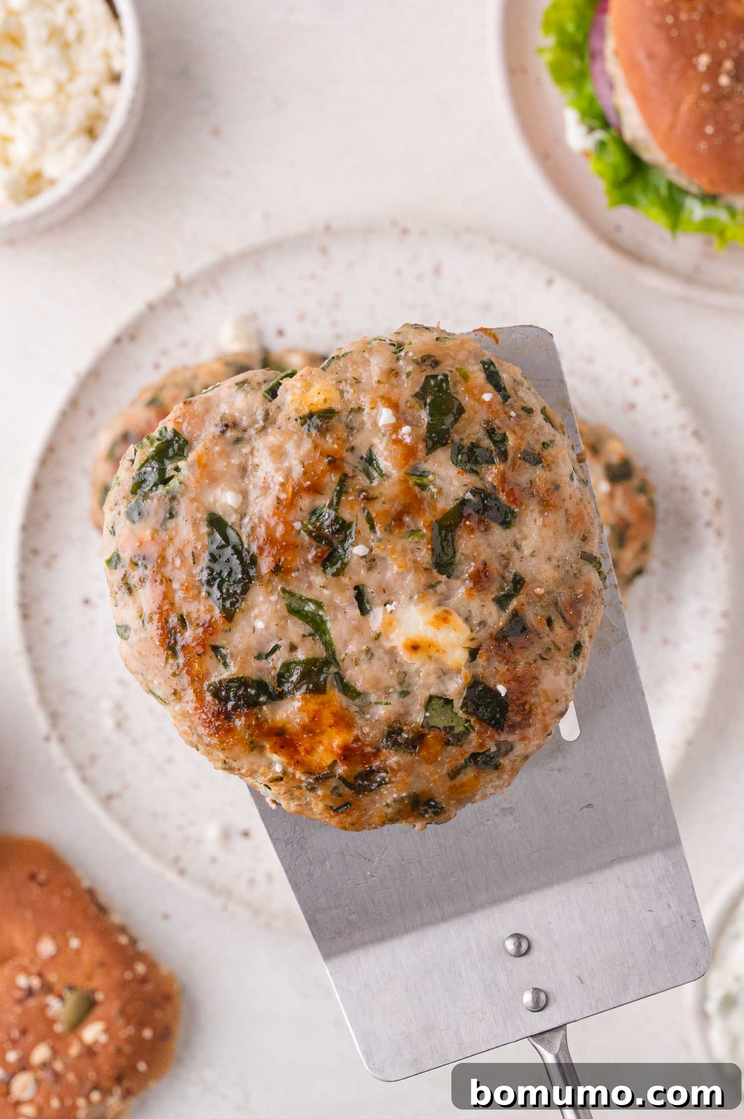 A cooked turkey burger patty with visible herbs and cheese is held on a metal spatula above a plate and burger buns.