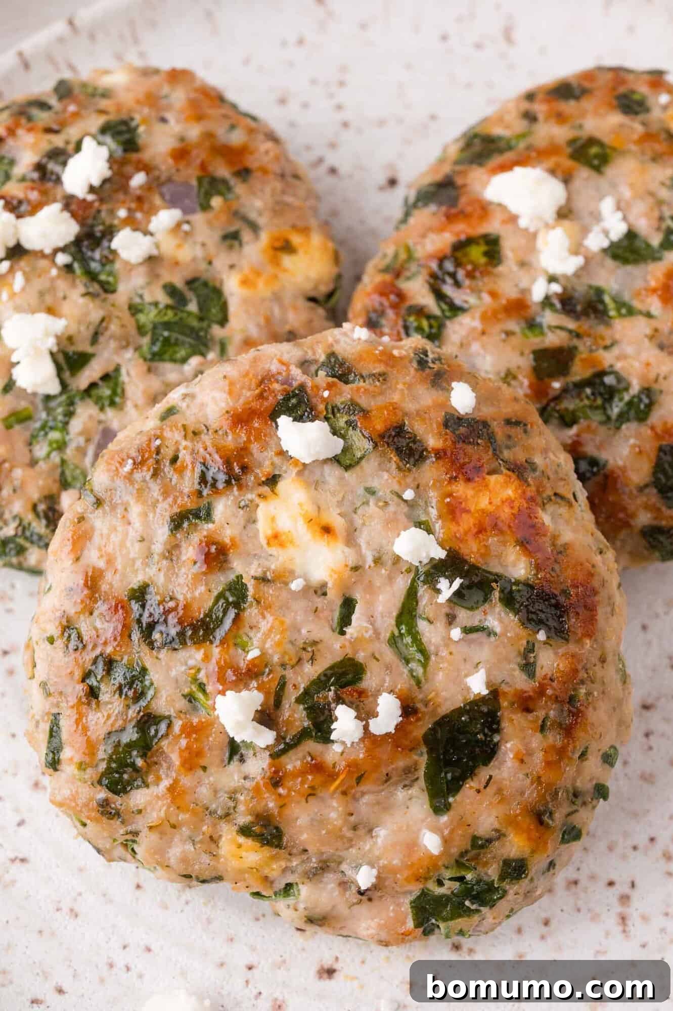 Three cooked greek turkey burgers mixed with spinach and feta cheese are shown on a speckled ceramic plate.
