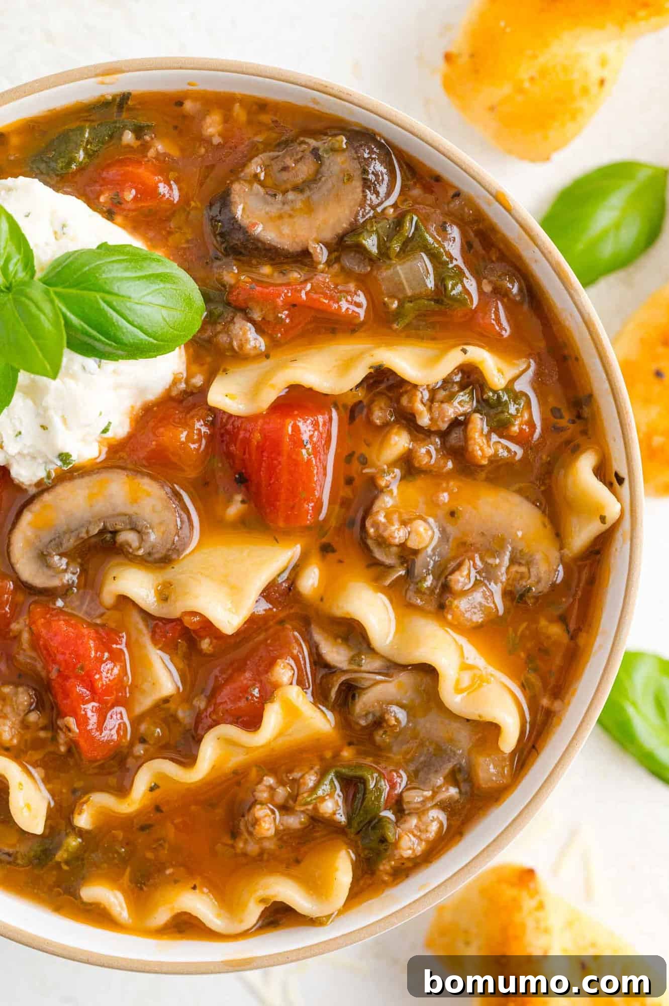 A beautifully presented bowl of lasagna soup, topped with a generous swirl of cheese and a sprig of fresh basil.