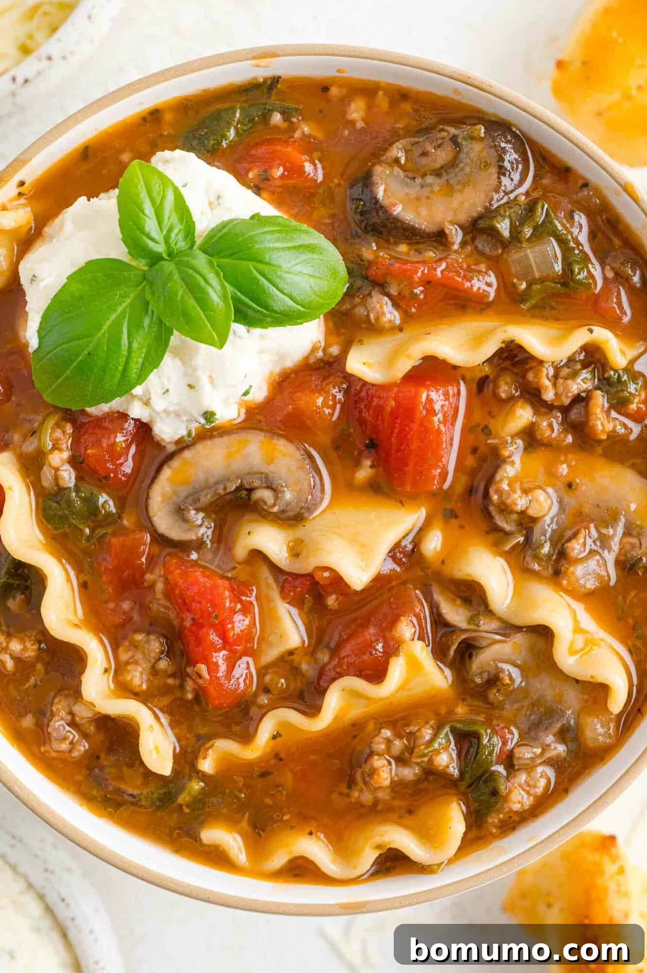 A bowl of rich lasagna soup garnished with fresh basil leaves and a dollop of creamy cheese.