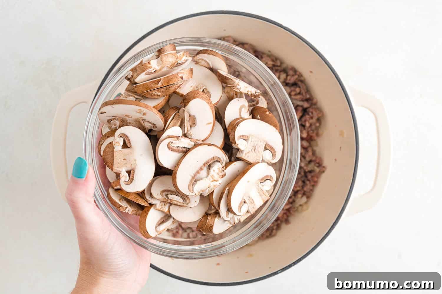 Fresh sliced mushrooms being added to the browned sausage and onions in a pan.