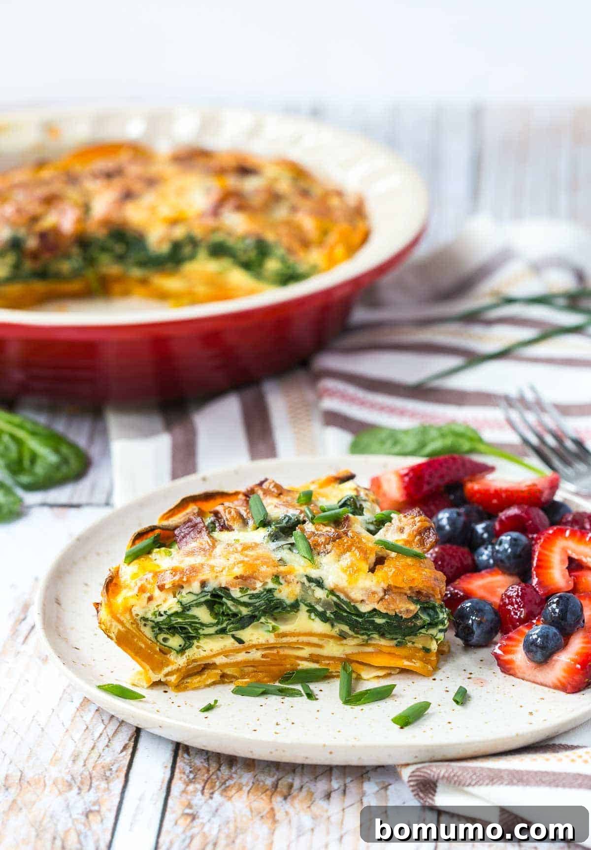 A slice of sweet potato crust quiche on a round white plate, garnished with fresh chives, accompanied by a side of fresh berries.