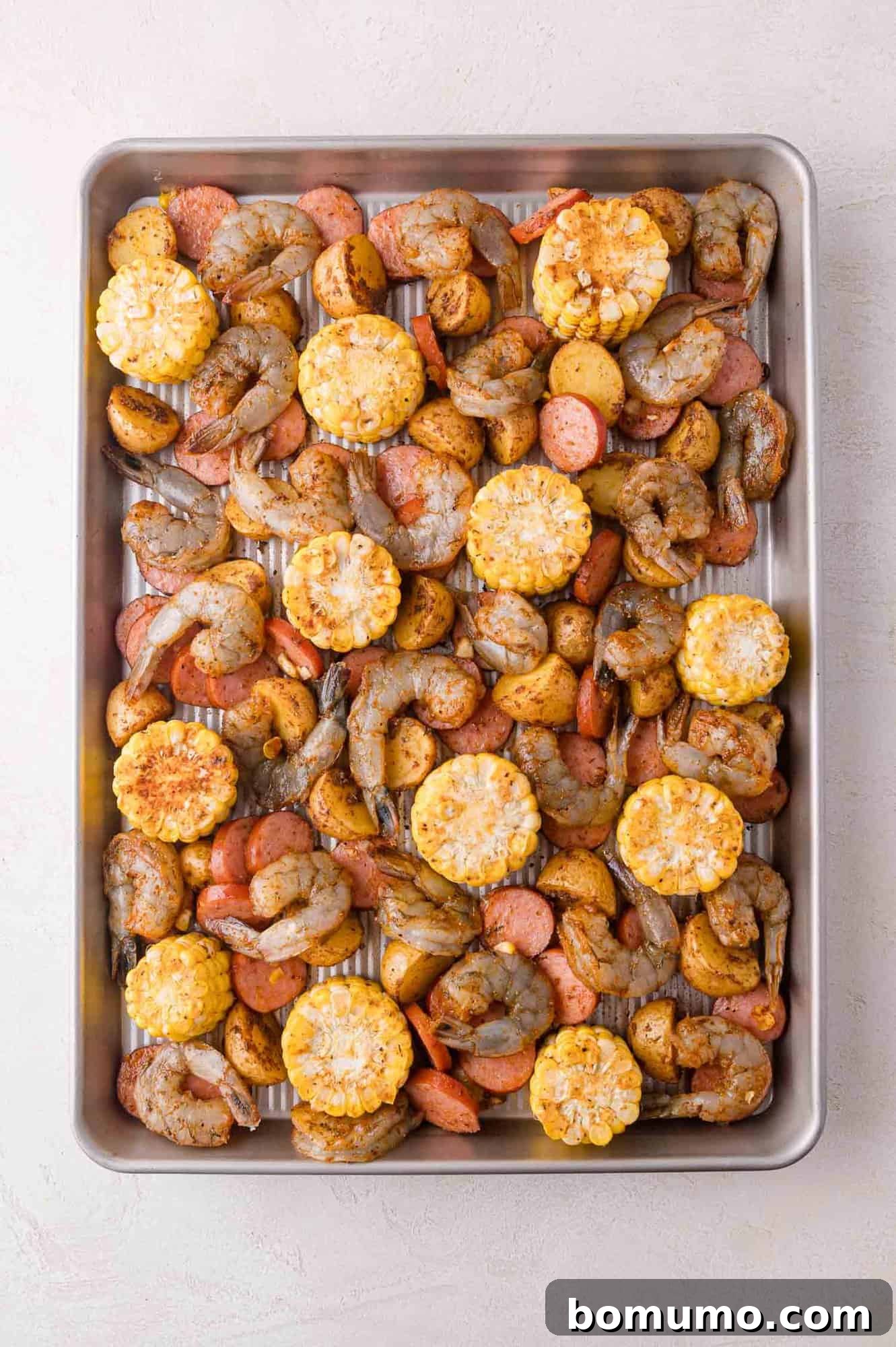 A sheet pan with raw shrimp, corn on the cob slices, sliced sausage, and diced potatoes seasoned with spices, ready to be cooked.