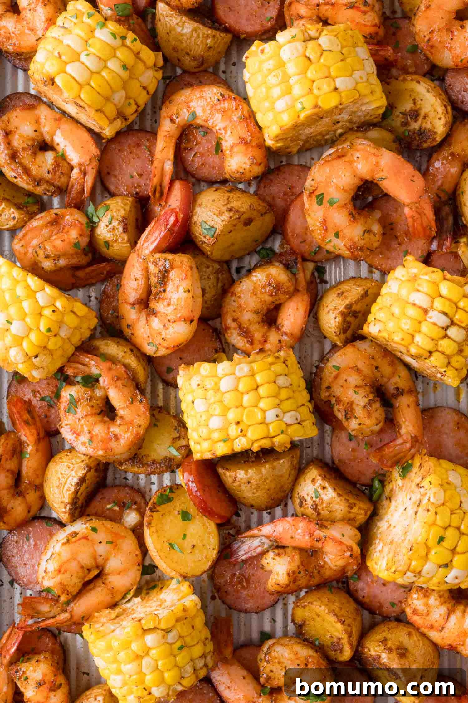 A sheet pan filled with roasted shrimp, sliced sausage, corn on the cob pieces, and halved baby potatoes, seasoned with herbs and spices.